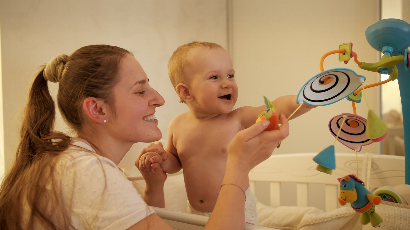 Portrait of smiling baby with mother playing with spinning toys hanging on mobile in bed at night